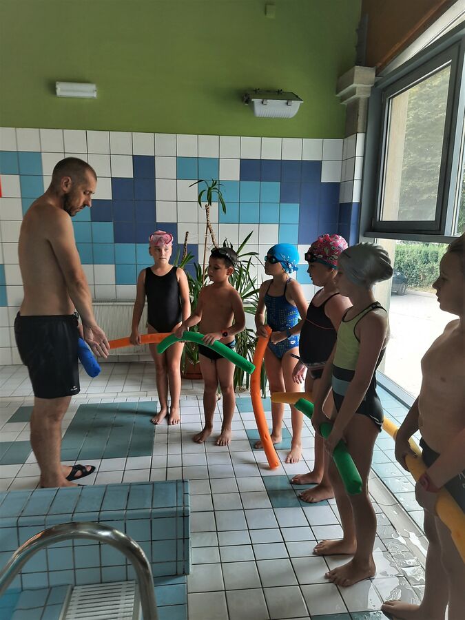 Zdjęcie przedstawia dzieci uczące się pływać. 6 dzieci stoi w rzędzie z instrumentami do nauki pływania. Przed nimi stoi instruktor. Dzieci stoją w skupieniu. W tle płytki oraz okno.