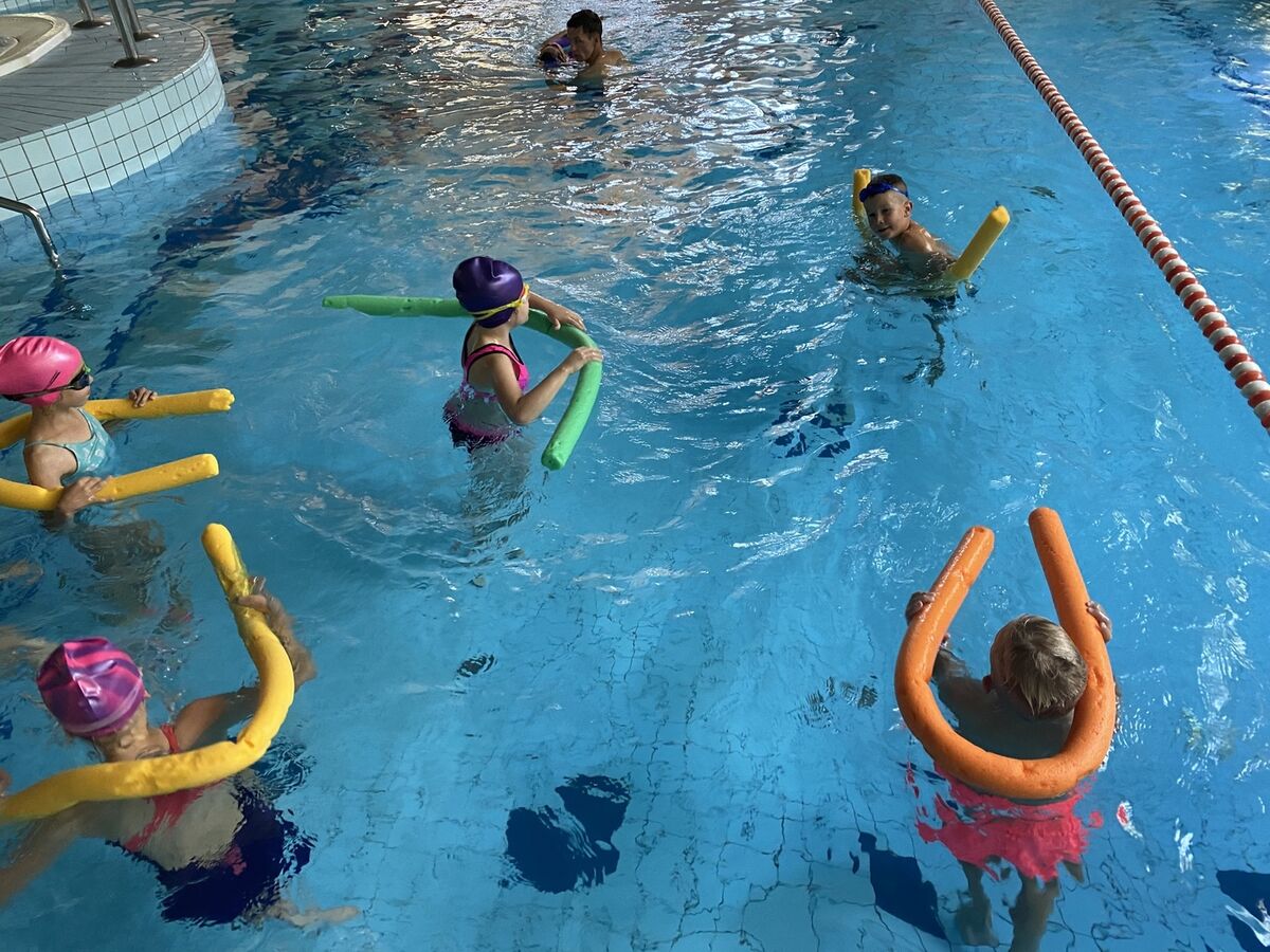 Zdjęcie przedstawia na pierwszym planie 5 dzieci, które uczą się pływać. Stoją w wodzie i w rękach mają sprzęt do pływania. 4 dzieci stoi bokiem, jedno stoi przodem i się uśmiecha. Na drugim planie instruktor uczy dziecko pływać. Dziecko płynie na plecach. 