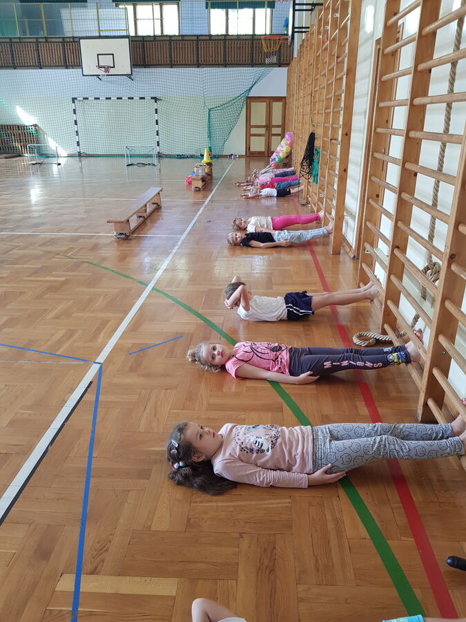 Zdjęcie przedstawia grupę dzieci. Dzieci leżą pod drabinkami. Mają nogi na pierwszym szczebelku. W tle sala gimnastyczna, kosz do koszykówki i bramka.