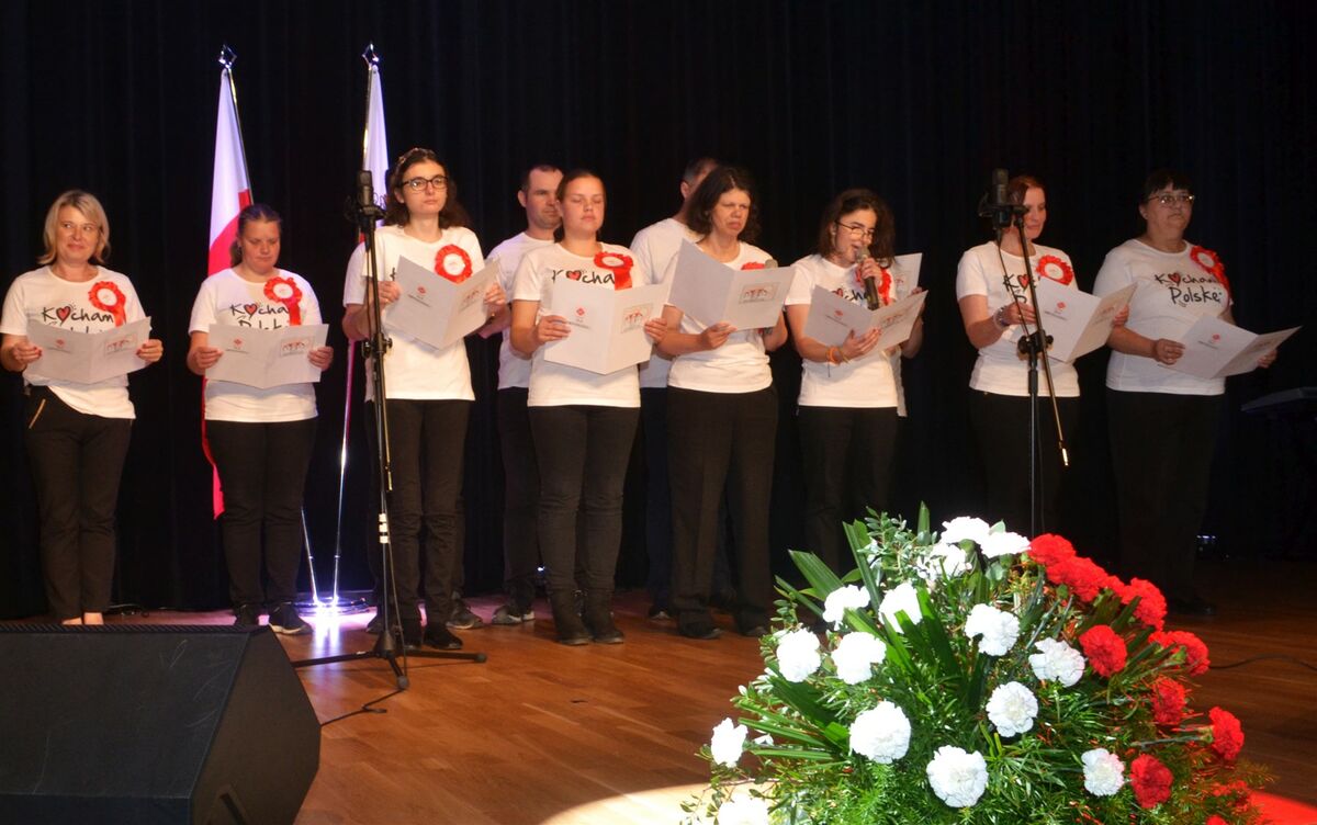 Uczestnicy Warsztatów terapii zajęciowej w Zembrzycach występują na scenie. Mają na sobie takie same koszulki z napisem Kocham Polskę. W rękach trzymają tekst piosenki.