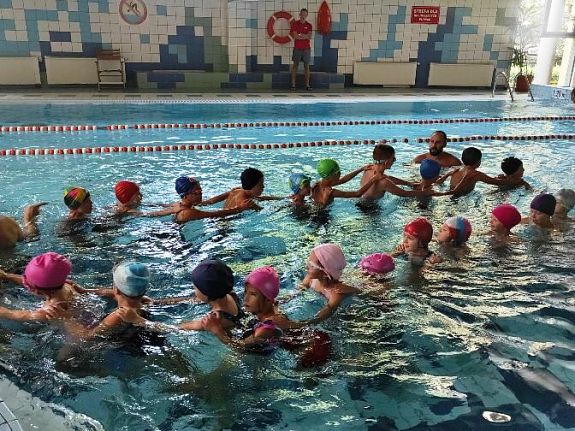 Zdjęcie przedstawia grupę dzieci, które trzymają się za ramiona. Obok nich stoi trzech instruktorów. Dzieci stoją w wodzie. W tyle druga część basenu - jest tam pusto, nikt nie pływa. Tory oddzielone są czerwonymi linami. W tle kryta pływalnia.